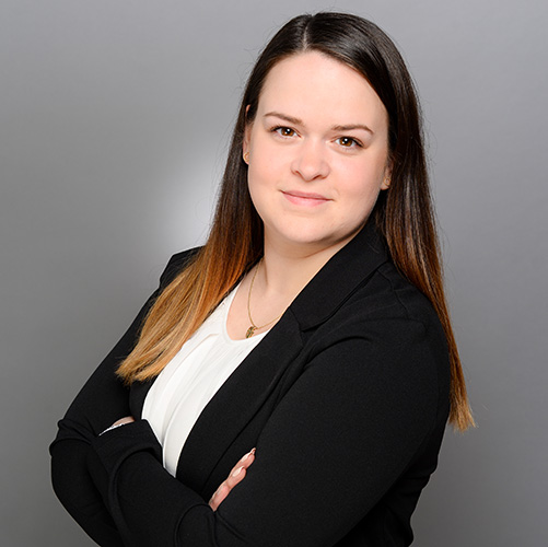 expertum - Eine Frau mit braunen Haaren im Blazer steht vor grauem Hintergrund, symbolisiert Fachkräftevermittlung in der Industrie.