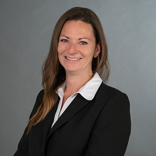 expertum - Eine Frau mit langen braunen Haaren im Business-Outfit vor grauem Hintergrund, steht für Industrie, Karriere und Zusammenarbeit.