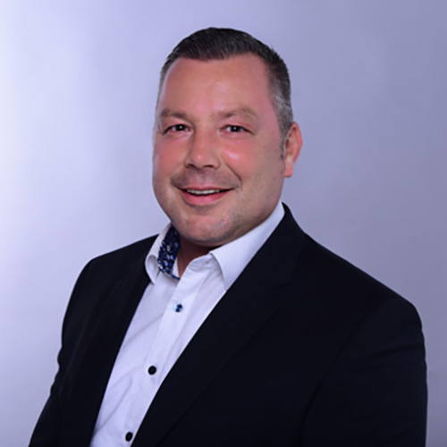 expertum - Ein Mann im dunklen Anzug und weißem Hemd blickt freundlich in die Kamera vor hellem Hintergrund - seriöses Auftreten im Arbeitskontext.