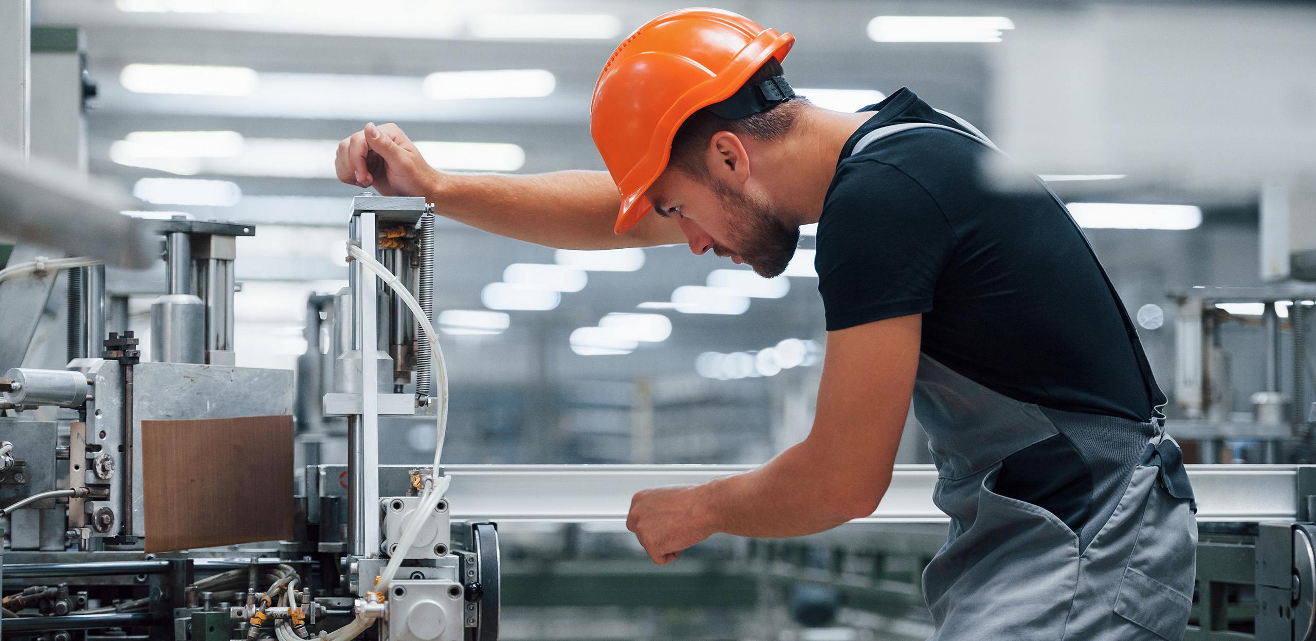 expertum - Ein Mann mit orangefarbenem Schutzhelm und grauem Overall bedient Maschinen in einer industriellen Produktionsumgebung. Er arbeitet konzentriert und justiert technische Komponenten – ein Beispiel für das fachliche Know-how und die Präzision, die qualifizierte Fachkräfte in Industrieprojekten sowie bei Personaldienstleistern wie expertum sowohl für Unternehmen als auch für Bewerber einbringen.