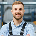 expertum - Ein Mann mit kurzem, hellbraunem Haar und Bart trägt ein hellblaues Hemd und eine dunkle Latzhose. Er steht lächelnd in einer industriellen oder werkstattähnlichen Umgebung. Das Bild zeigt eine typische Arbeitssituation im handwerklichen oder industriellen Bereich und passt damit zum Kontext der Personalvermittlung, der Fachkräftegewinnung und der Zusammenarbeit zwischen Bewerbern und Unternehmen in der Industrie.