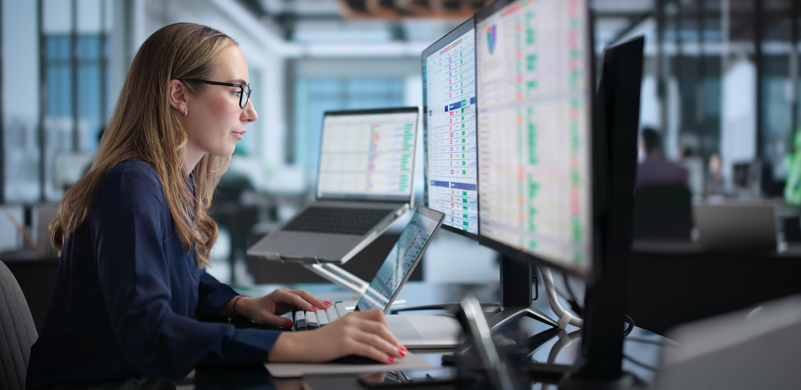 expertum - Eine Frau mit Brille arbeitet konzentriert an einem Schreibtisch in einem modernen Büro, umgeben von mehreren großen Computerbildschirmen mit farbigen Datendiagrammen. Die Szene vermittelt einen Einblick in die Arbeitswelt von Fachkräften und zeigt Tätigkeiten wie Datenanalyse oder Projektarbeit, wie sie im industriellen Umfeld und bei Personaldienstleistern für Unternehmen und Bewerber relevant sind.