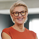 expertum - Eine ältere, freundlich blickende Frau mit kurzen blonden Haaren, Brille und orangefarbenem Oberteil steht in einem Innenraum vor einem unscharfen Hintergrund. Sie strahlt Kompetenz und Erfahrung aus – Eigenschaften, die sowohl für Bewerberinnen und Bewerber als auch für Unternehmen in der modernen Industrie- und Arbeitswelt im Bereich Personalvermittlung und Projektarbeit von Bedeutung sind.