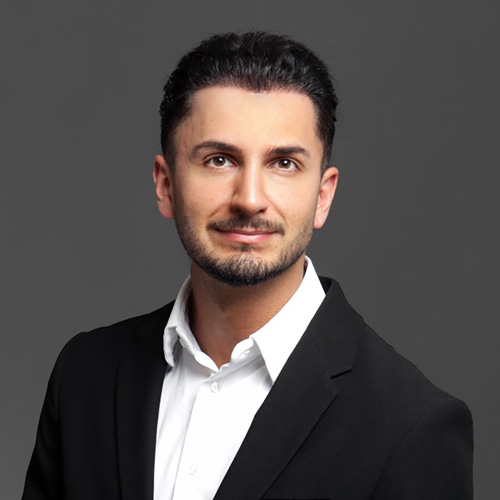 expertum - Ein Mann mit kurzem dunklem Haar und Bart im Business-Outfit vor grauem Hintergrund, symbolisiert Personalvermittlung in der Industrie.