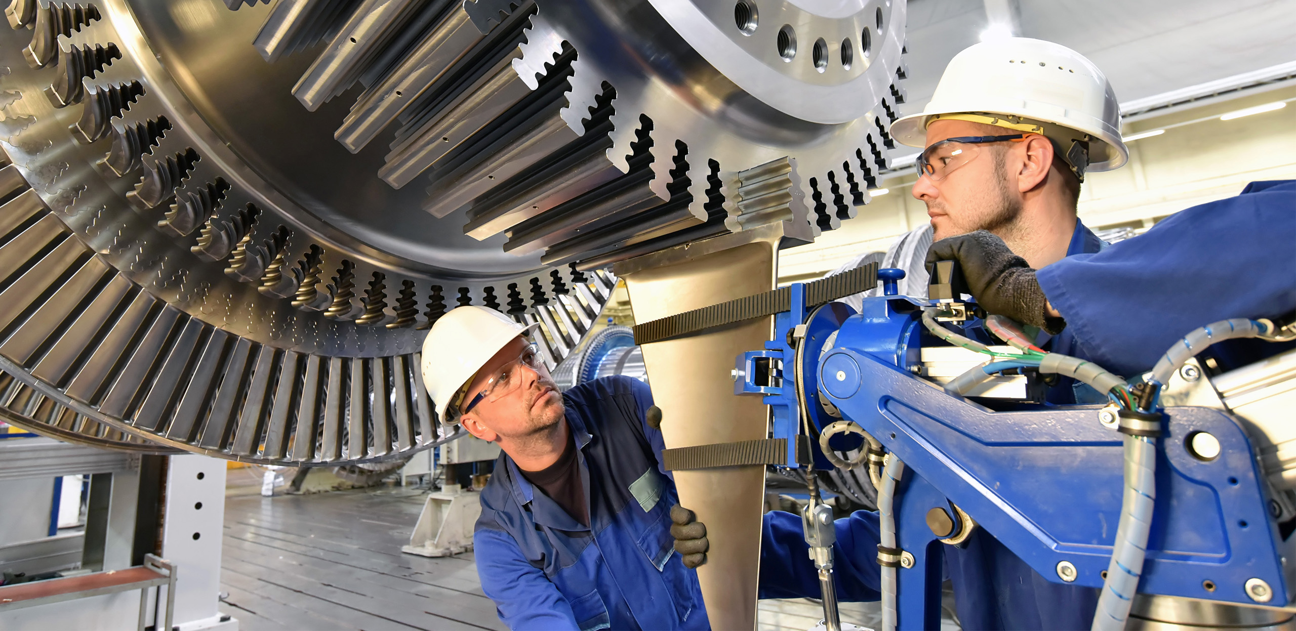 expertum - Zwei Ingenieure mit Helmen und blauer Arbeitskleidung prüfen und montieren in einem modernen Werk Teile einer großen Industrieturbine mithilfe spezieller Geräte und Werkzeuge. Die Szene zeigt die praxisnahe Zusammenarbeit von Fachkräften in der Industrie, wie sie für den beruflichen Alltag, die Projektarbeit sowie für die Vermittlung von Personal zwischen Unternehmen und technischen Experten typisch ist.
